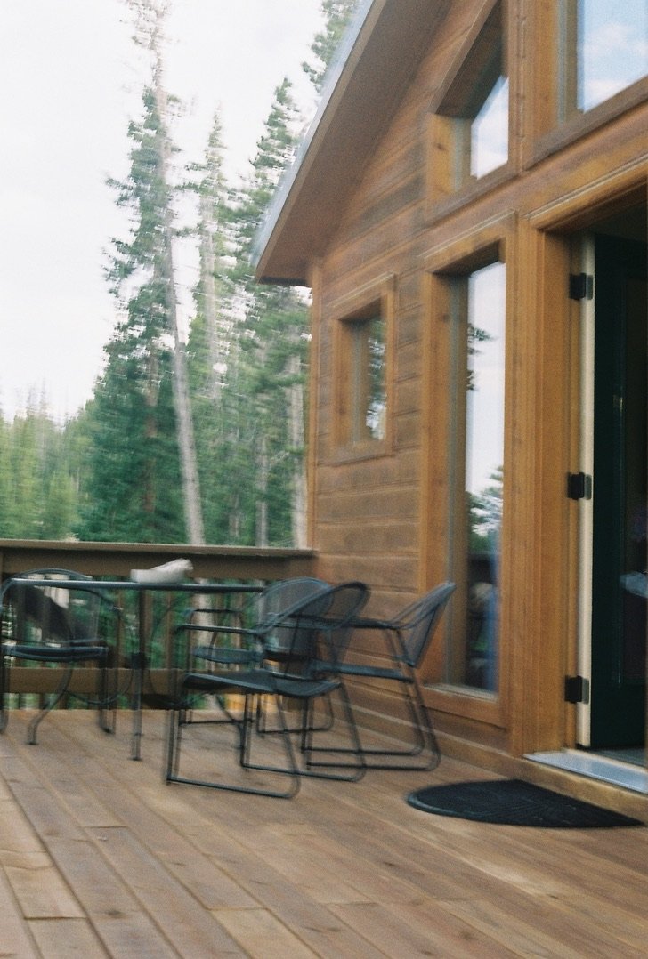Wooden outdoor deck with black metal chairs and a table, attached to a wooden cabin with large windows, surrounded by trees.