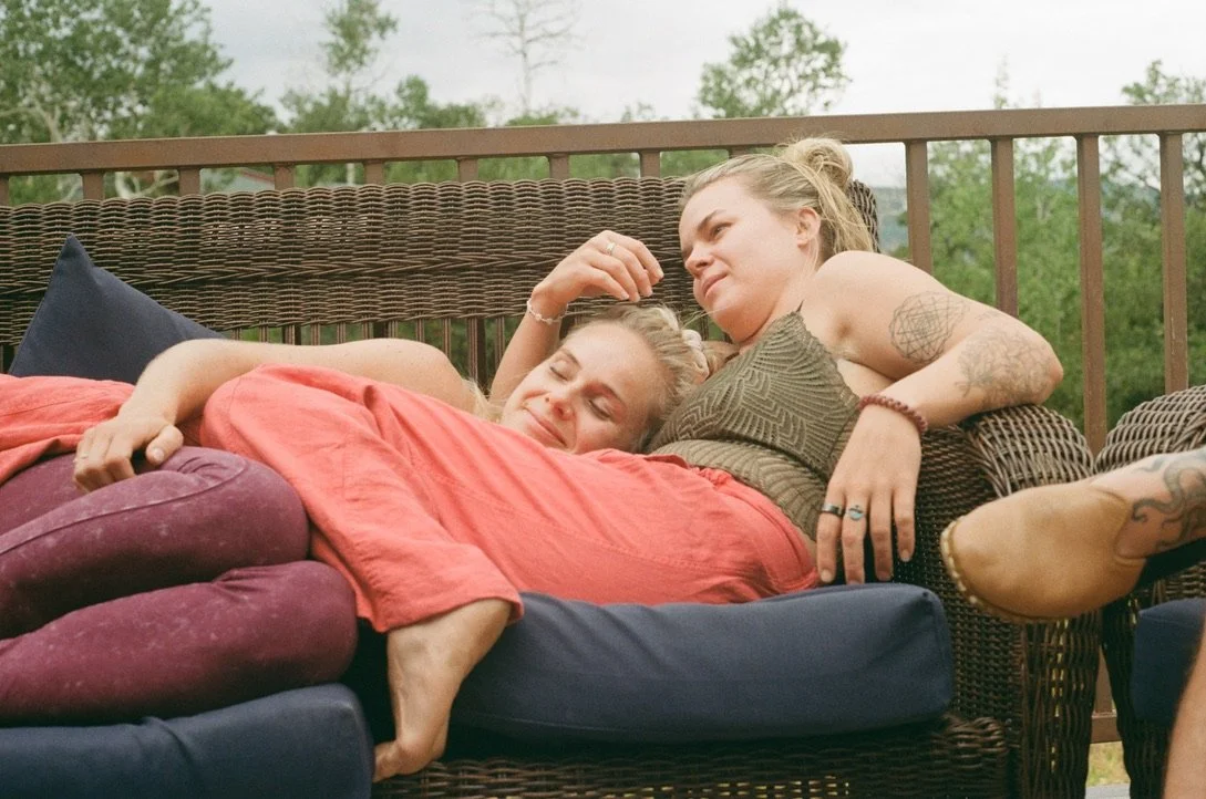Two women relaxing on a wicker outdoor couch, one lying down with a red blanket and the other sitting beside her, looking at her with a smile, surrounded by green trees.