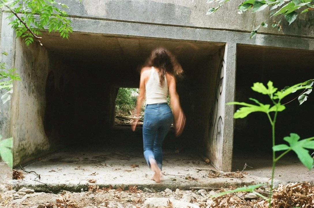 A woman walking barefoot through a tunnel under a concrete structure surrounded by greenery.