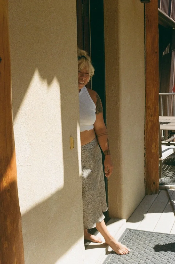 A woman with blonde hair smiling and peeking out from behind a wall on a porch, wearing a white crop top and patterned pants, barefoot in sunlight.