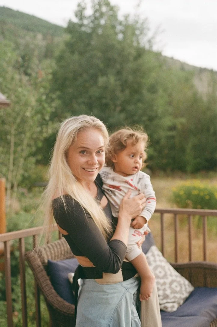 A woman with long blonde hair smiling while holding a young child with curly hair on an outdoor wooden deck, with green trees and a mountain in the background.