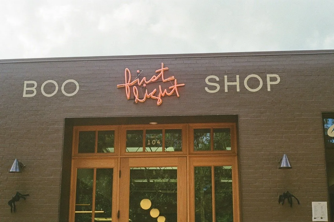 Storefront with sign reading 'BOO', pink neon sign saying 'first light', and 'SHOP' on the wall, with wooden doors at the entrance.