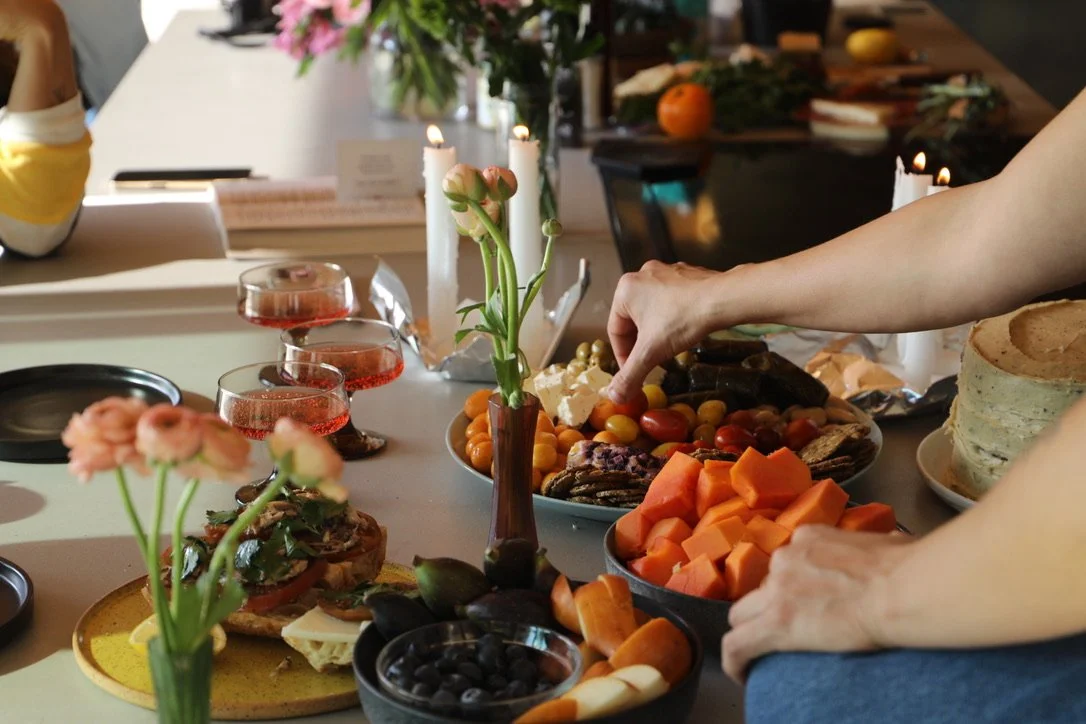 A table set with various foods including sliced vegetables, cheeses, fruits, and desserts, with hands reaching for the food, and lit candles in the background.