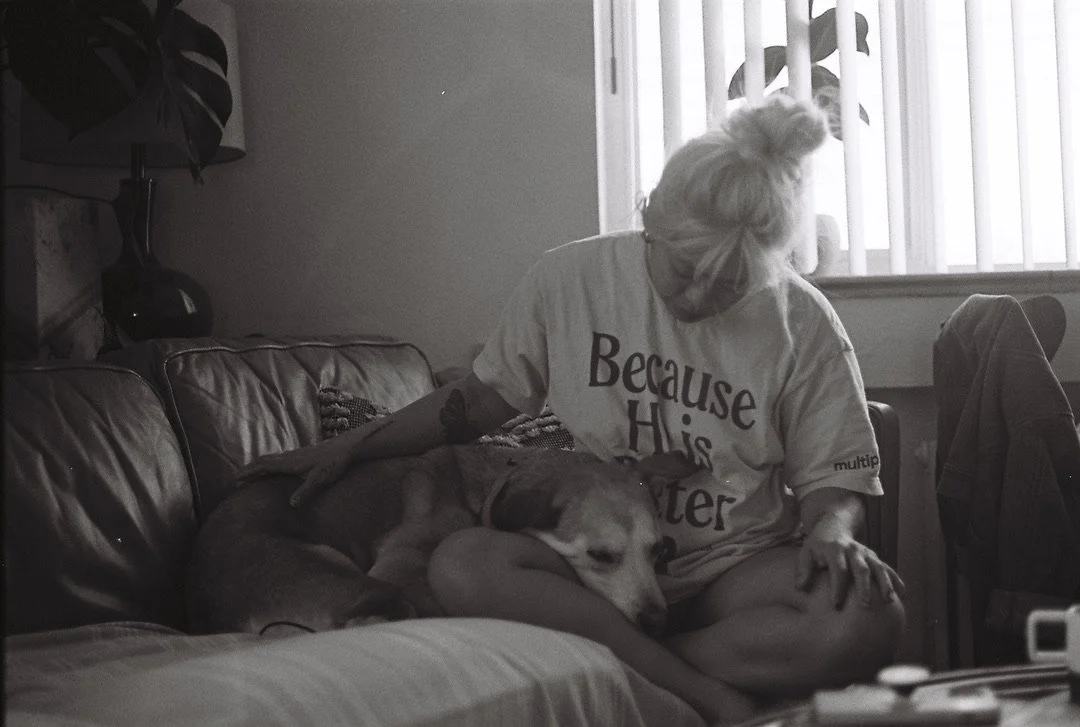 A woman with light-colored hair in a bun sitting on a couch, petting a large dog resting on her lap, in a room with a window and blinds.