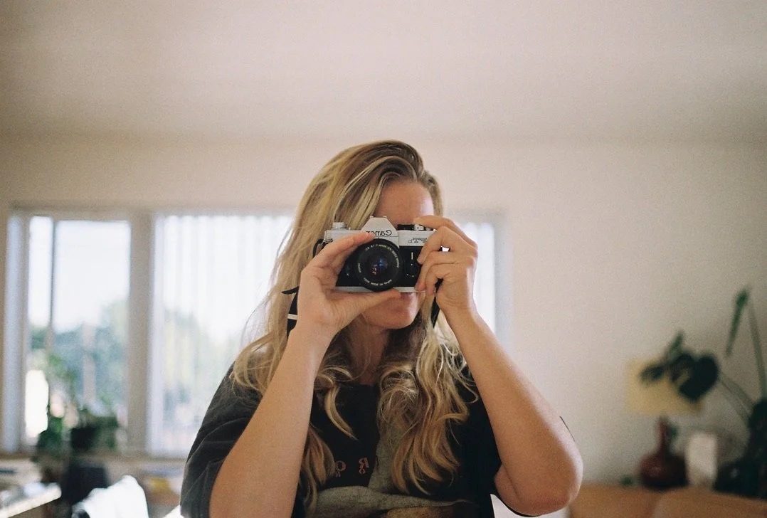 Woman with long wavy blonde hair taking a photo with a camera in a room with large windows and plants.