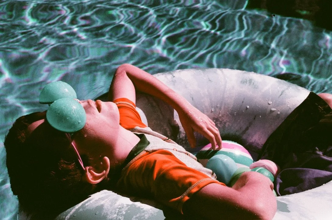A person floating on a pool float with sunglasses resting on their forehead, surrounded by swimming pool water and colorful balls.