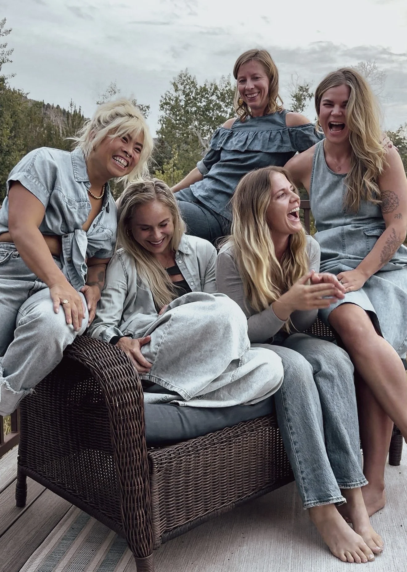 Group of six women laughing and enjoying time together outdoors on a patio, sitting and standing around a wicker couch.