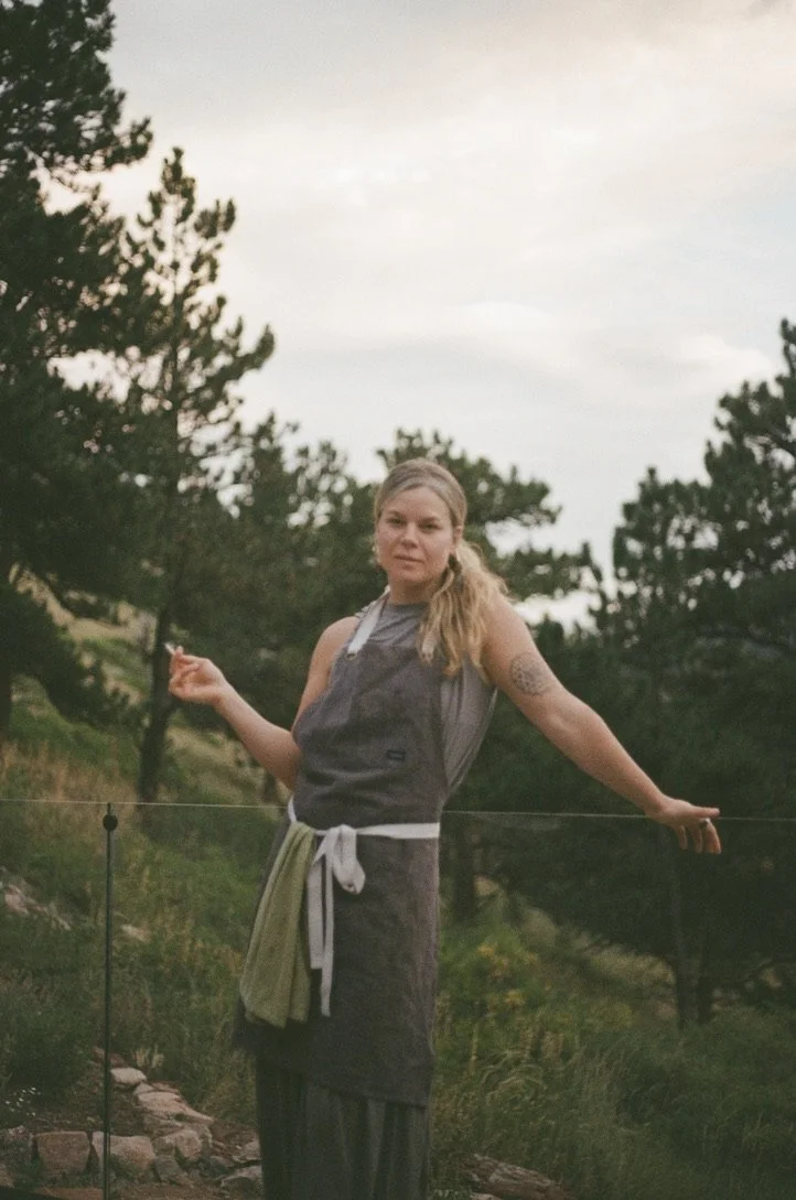 A woman with blonde hair standing outdoors with trees in the background, wearing a sleeveless shirt and an apron, holding a towel, with a slight smile.