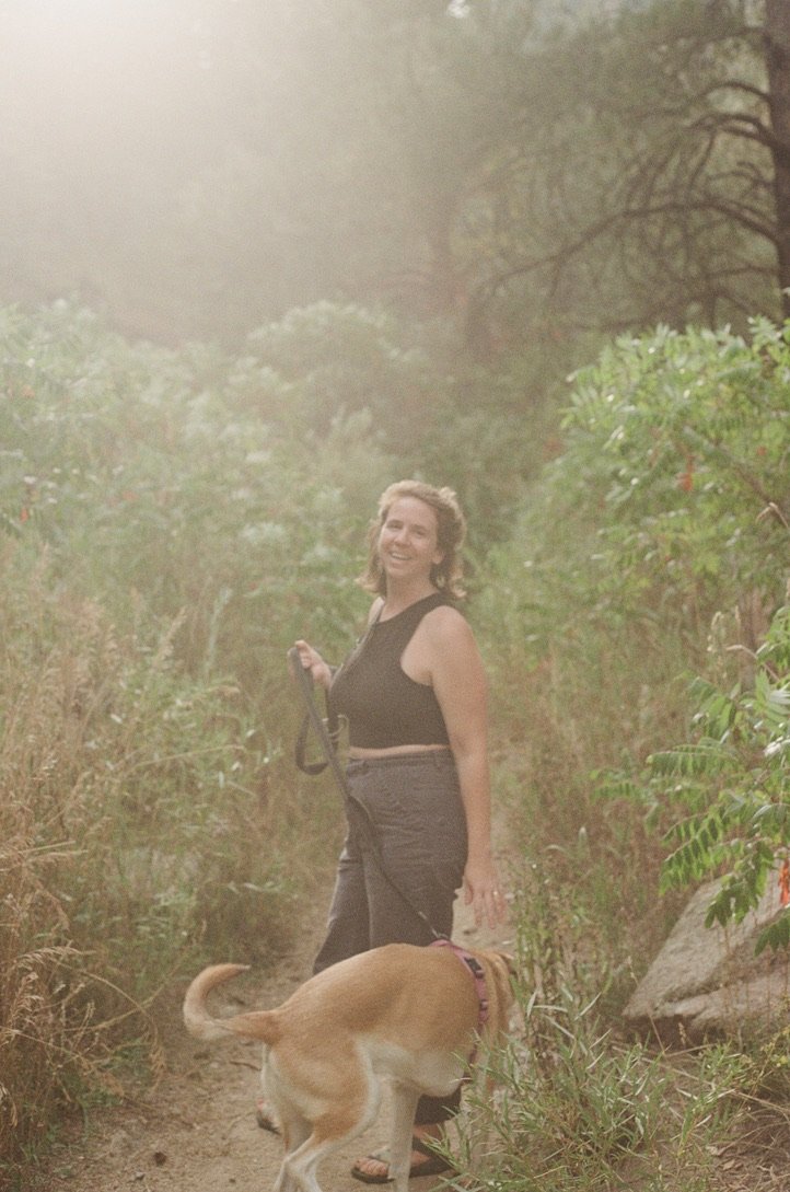 A woman smiling while walking her dog on a nature trail surrounded by green bushes and trees.