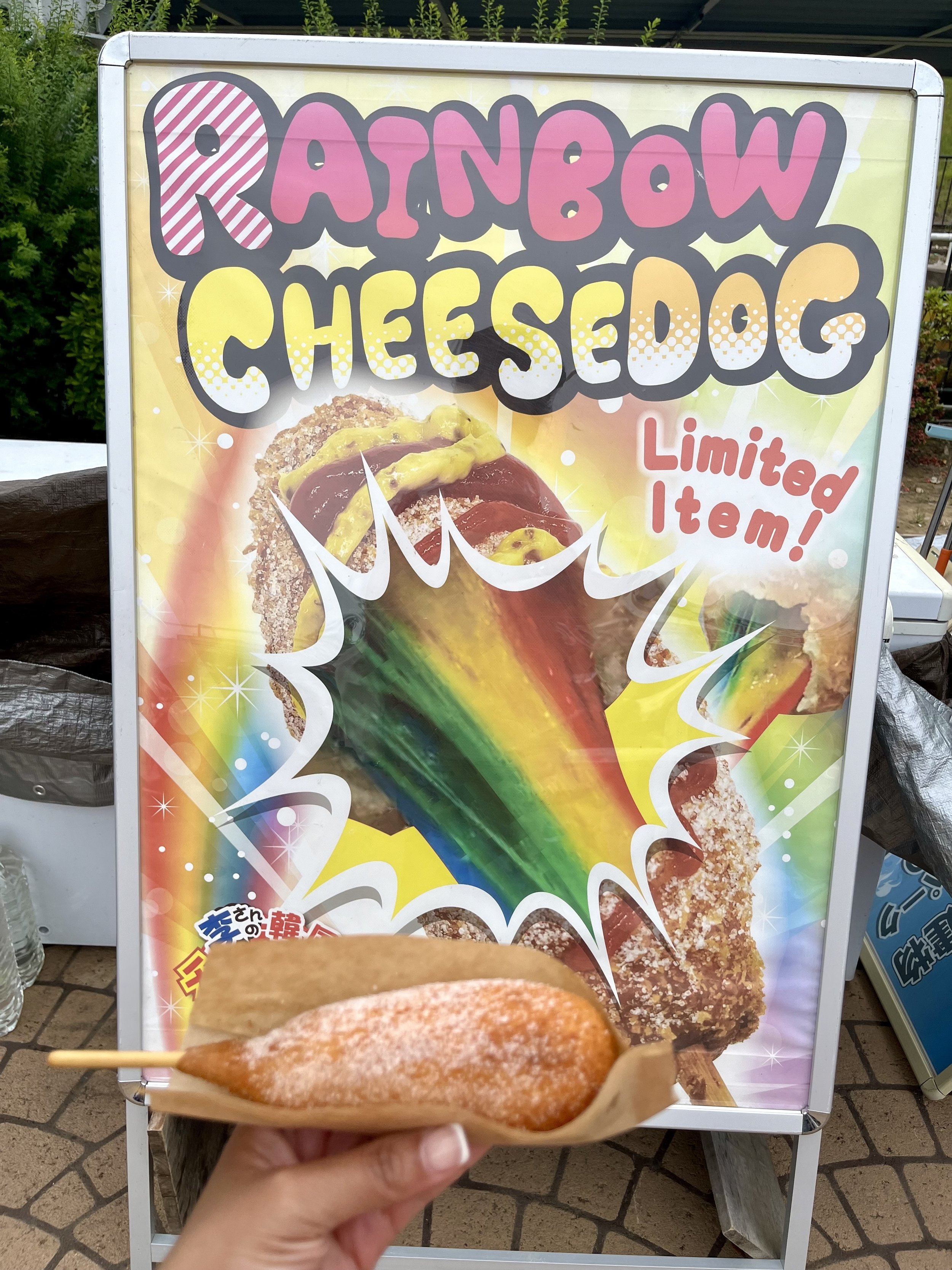 Rainbow Cheese Dog sold at Yomiuriland as a limited time themed snack. The colorful corn dog is displayed in front of its menu sign and is one of the park's unique food offerings.
