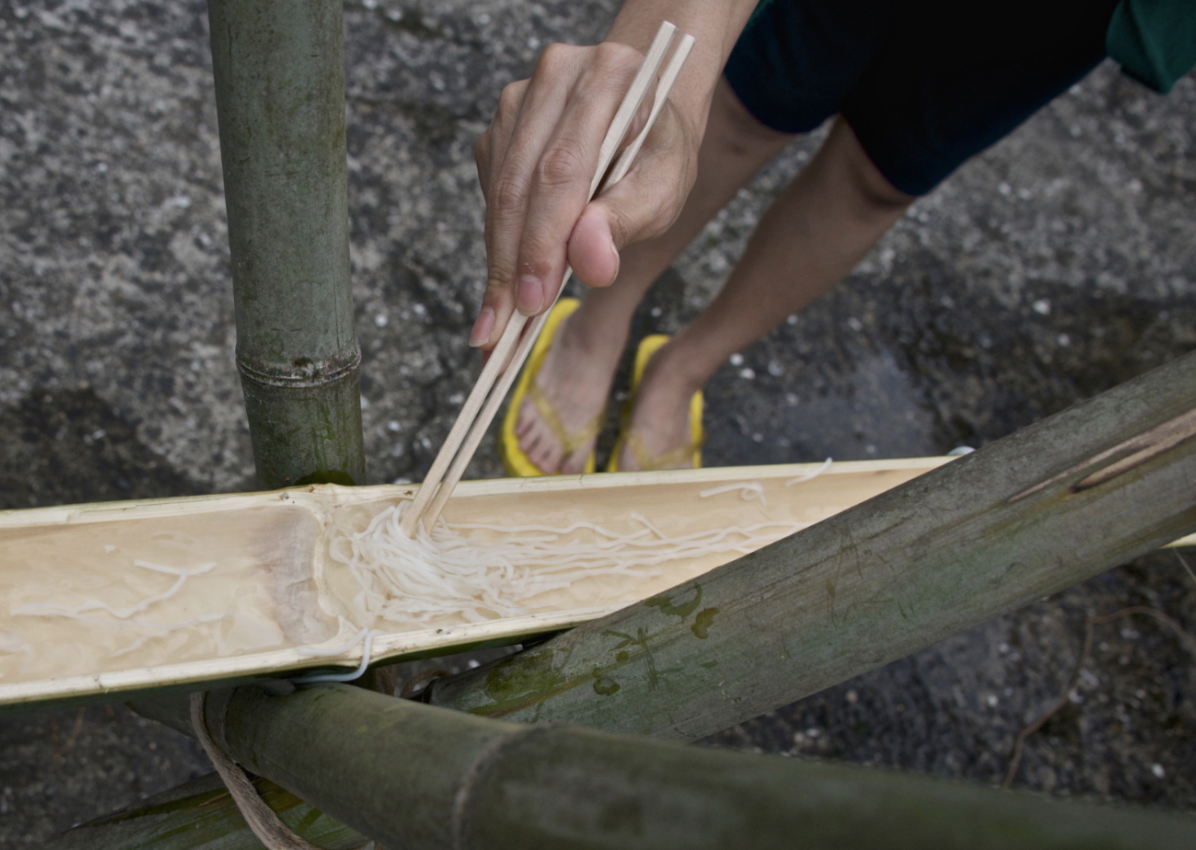 A cropped image of a person's hand holding wooden chopsticks grabs noodles from a bamboo shoot split halfway open.