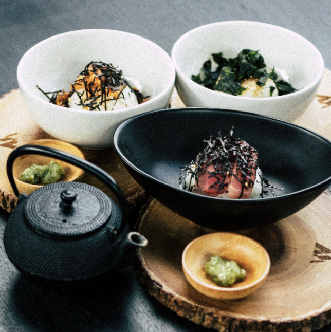 A set meal with 3 main bowls of food, rice fish, and a vegetable, 2 small bowls of paste, and a black iron tea kettle.