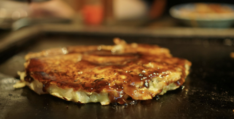 A Japanese-style circular omelette with a brown soy sauce glaze on top is grilled on a flat sheet.