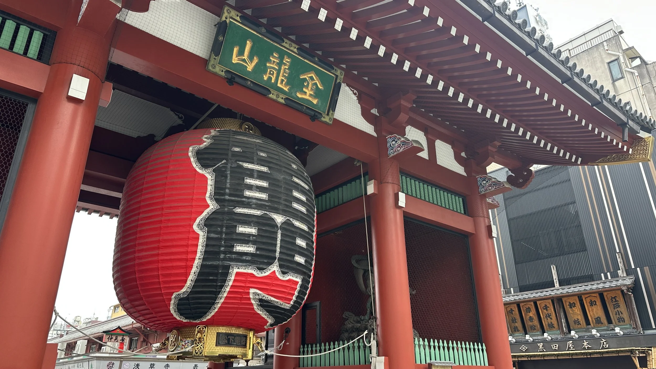 Red lantern hangs from a red and green gate at the front of a shopping street in front of a shrine in Senso-ji in the Asakusa area of Tokyo, Japan.
