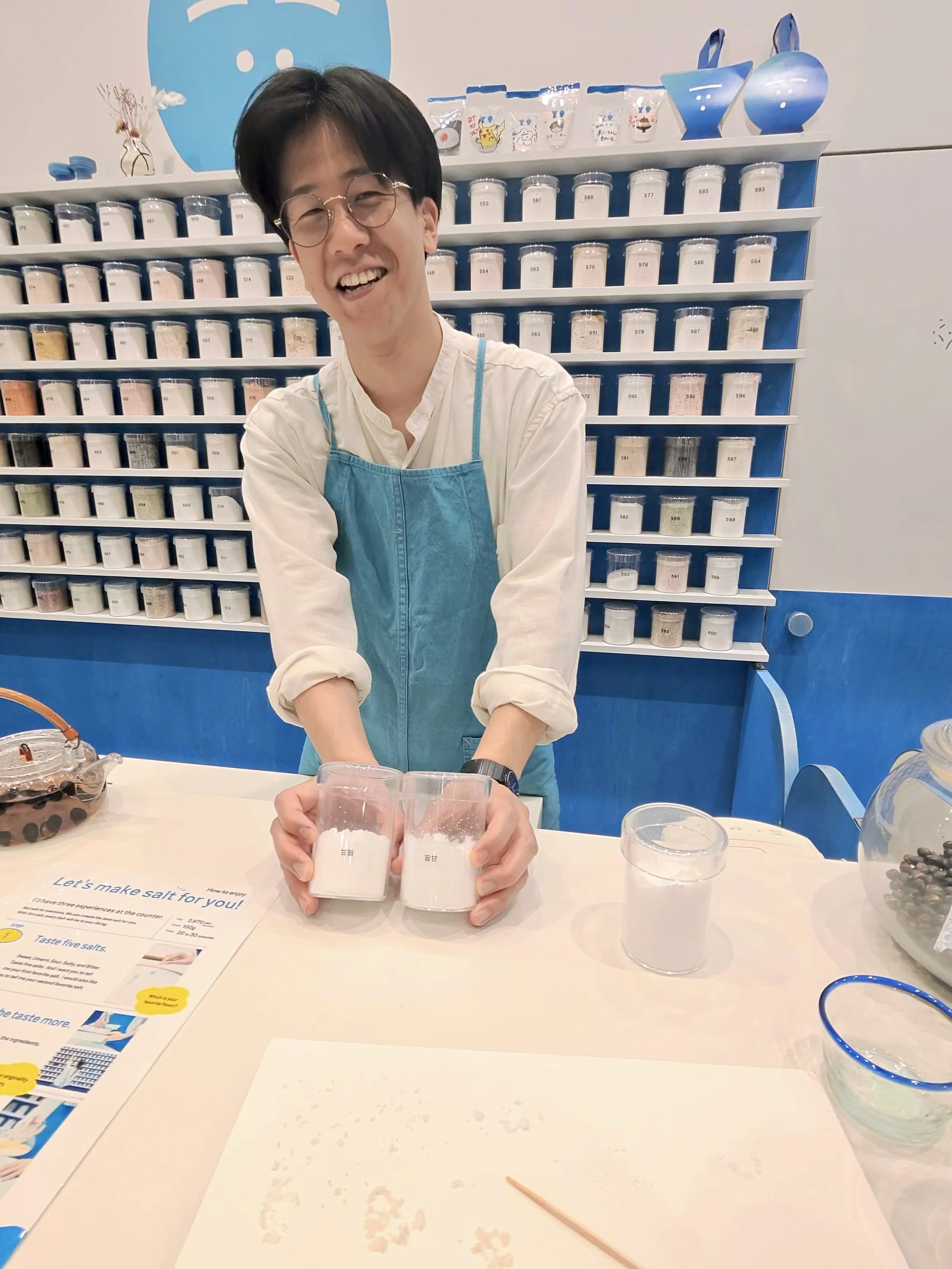 Staff member preparing a custom salt blend during the Guruguru Shakashaka salt making experience in Tokyo. Shelves of different salts are displayed behind the counter as the personalized blend is prepared.