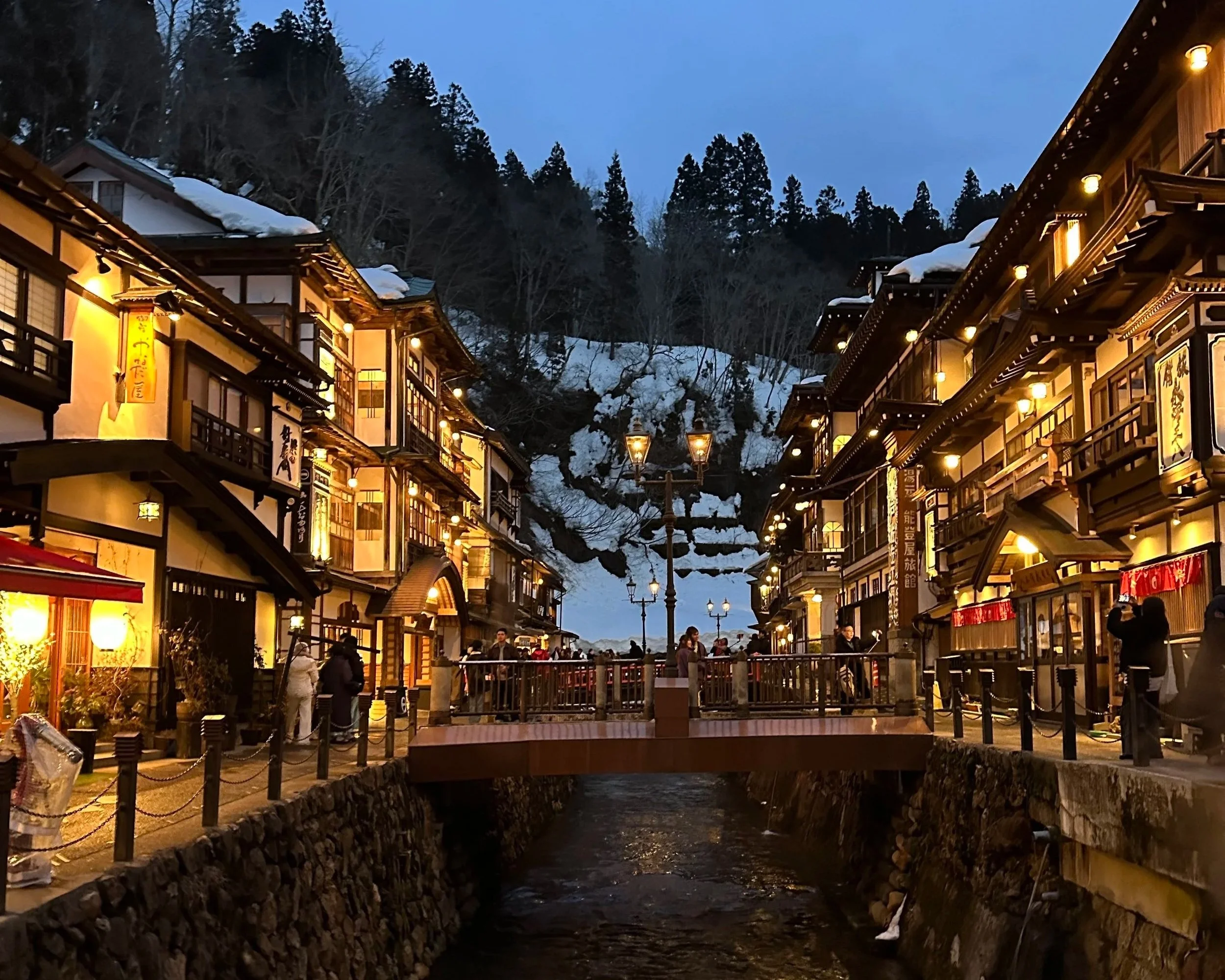 An old onsen village nestled in the snow capped mountains with light colored 3 story buildings with dark trimming and a river running through the middle of the image.