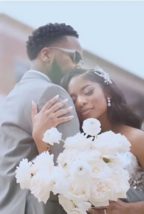 A bride and groom embrace at their wedding, with the bride holding a large bouquet of white flowers and the groom wearing sunglasses.
