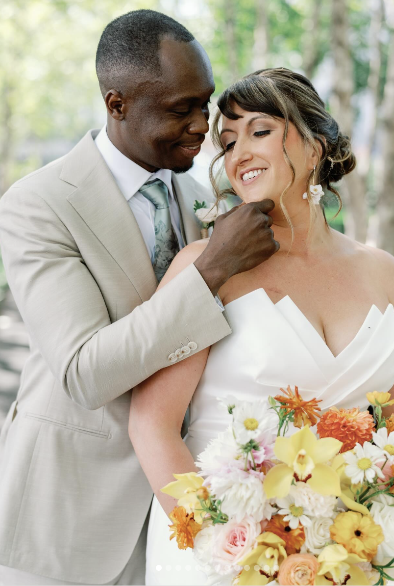 A groom with dark skin gently touching a bride's chin, both smiling with eyes closed, outdoors with greenery, a wedding bouquet with orange, yellow, pink, and white flowers.