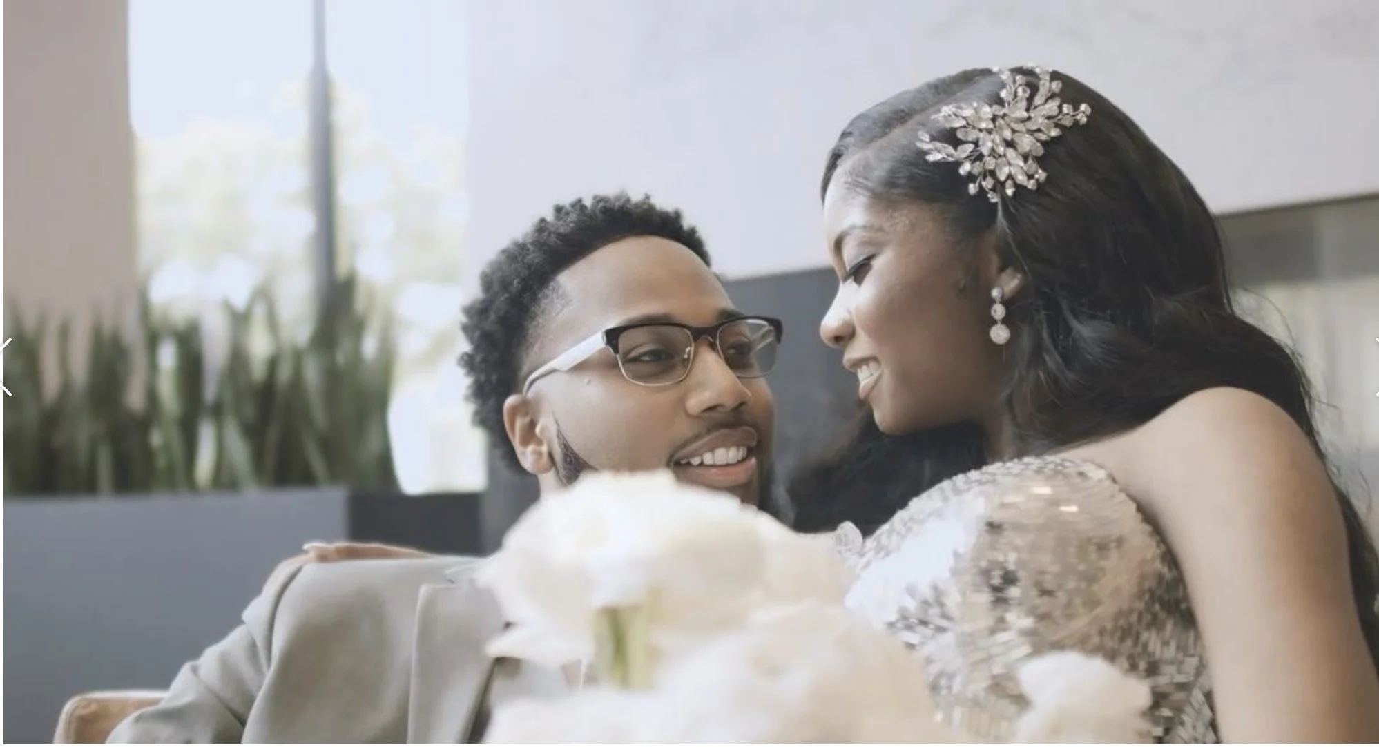 A joyful couple is embracing, smiling, and sharing a close moment indoors with a flower in the foreground, celebrating a special occasion.