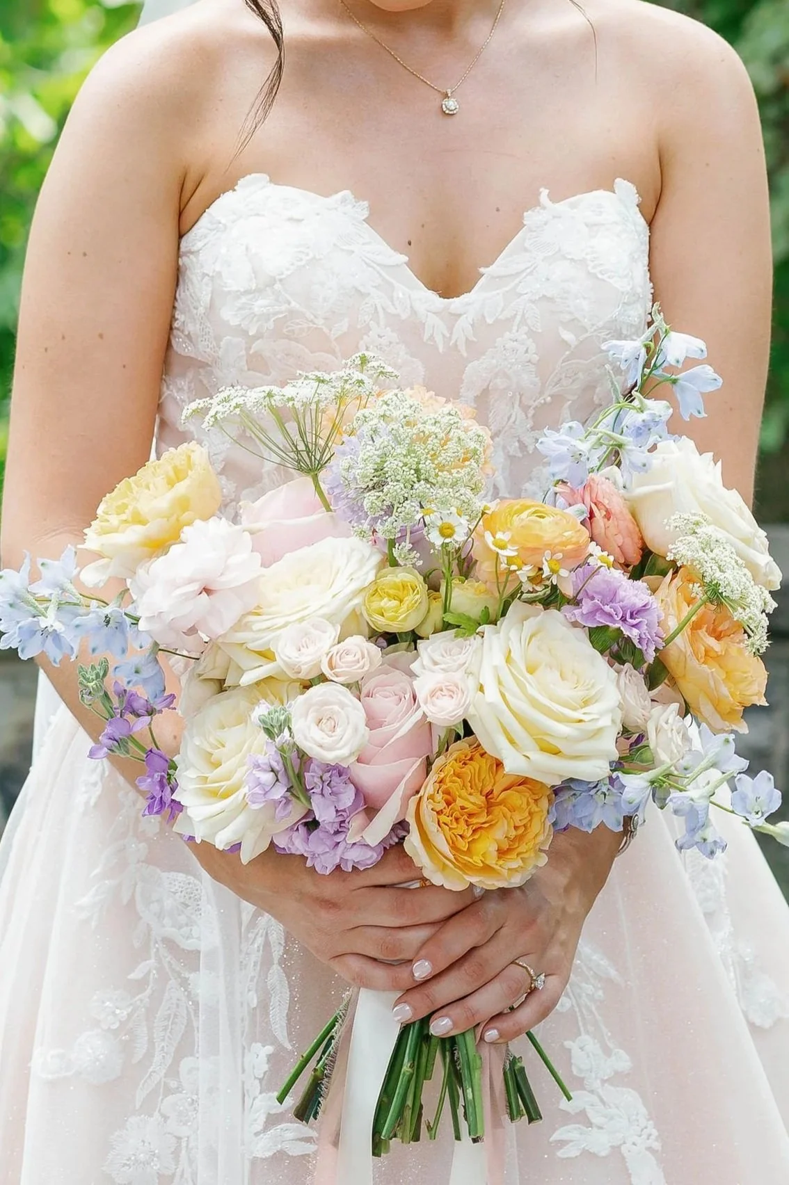 A bride in a lace wedding dress holding a large bouquet of pastel and white flowers, with greenery in the background.