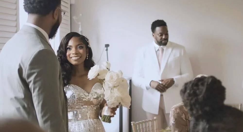 A bride holding a bouquet of white flowers, smiling at a groom in a light-colored suit, with a man in a white suit observing them during a wedding celebration.