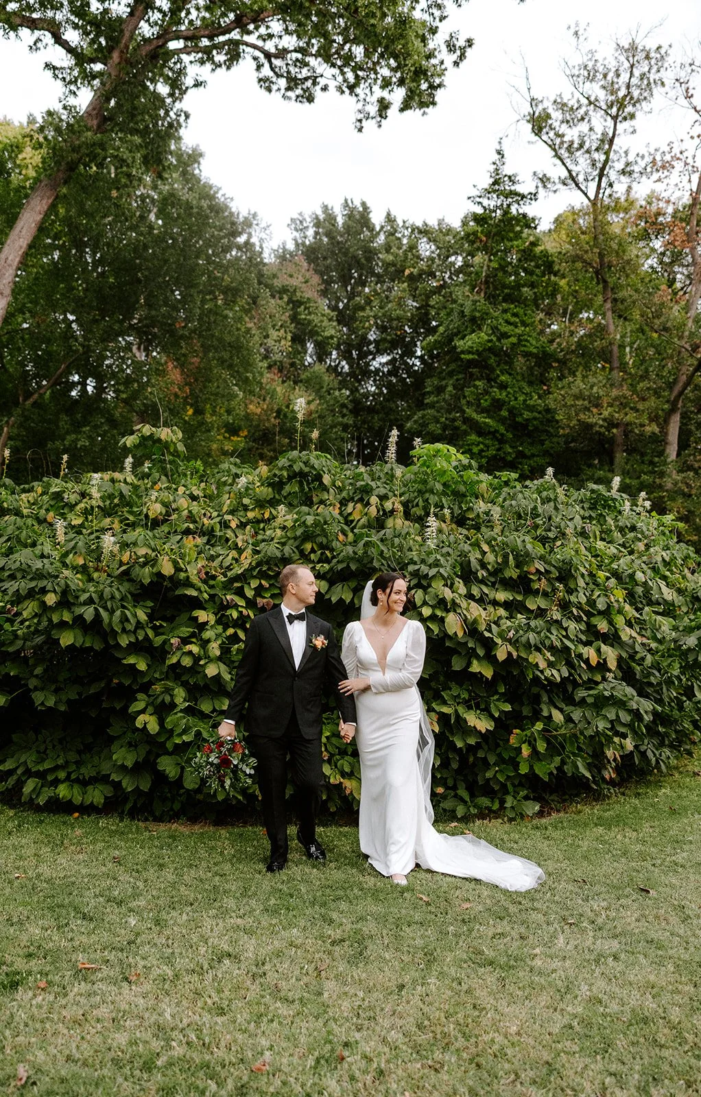 A bride and groom walking arm in arm outdoors near a dense green shrub, with trees in the background. The bride is wearing a white wedding gown and veil, holding a bouquet, and the groom is in a black tuxedo with a bow tie.