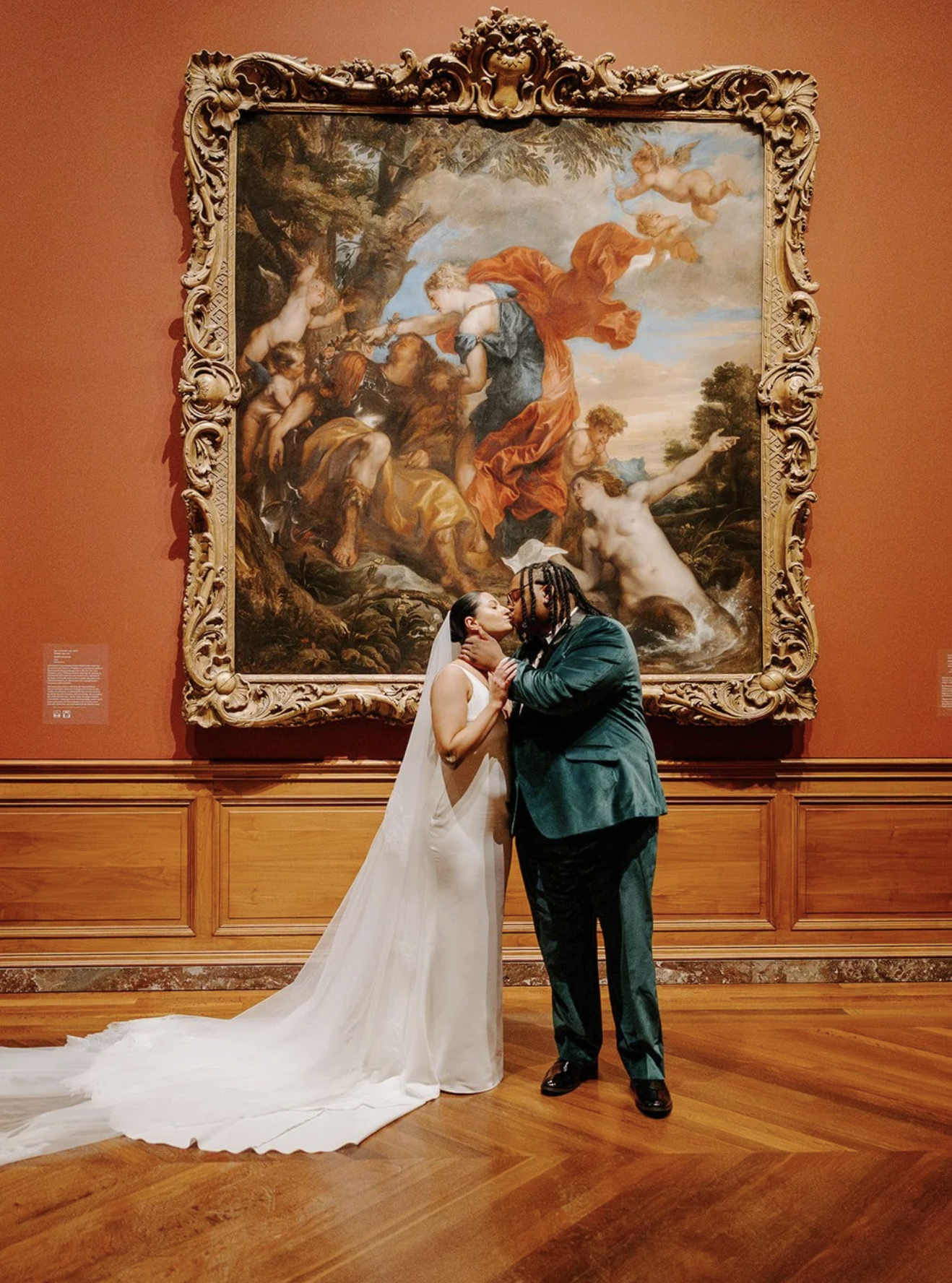 A bride and groom kiss in front of a large ornate framed painting in an art gallery.