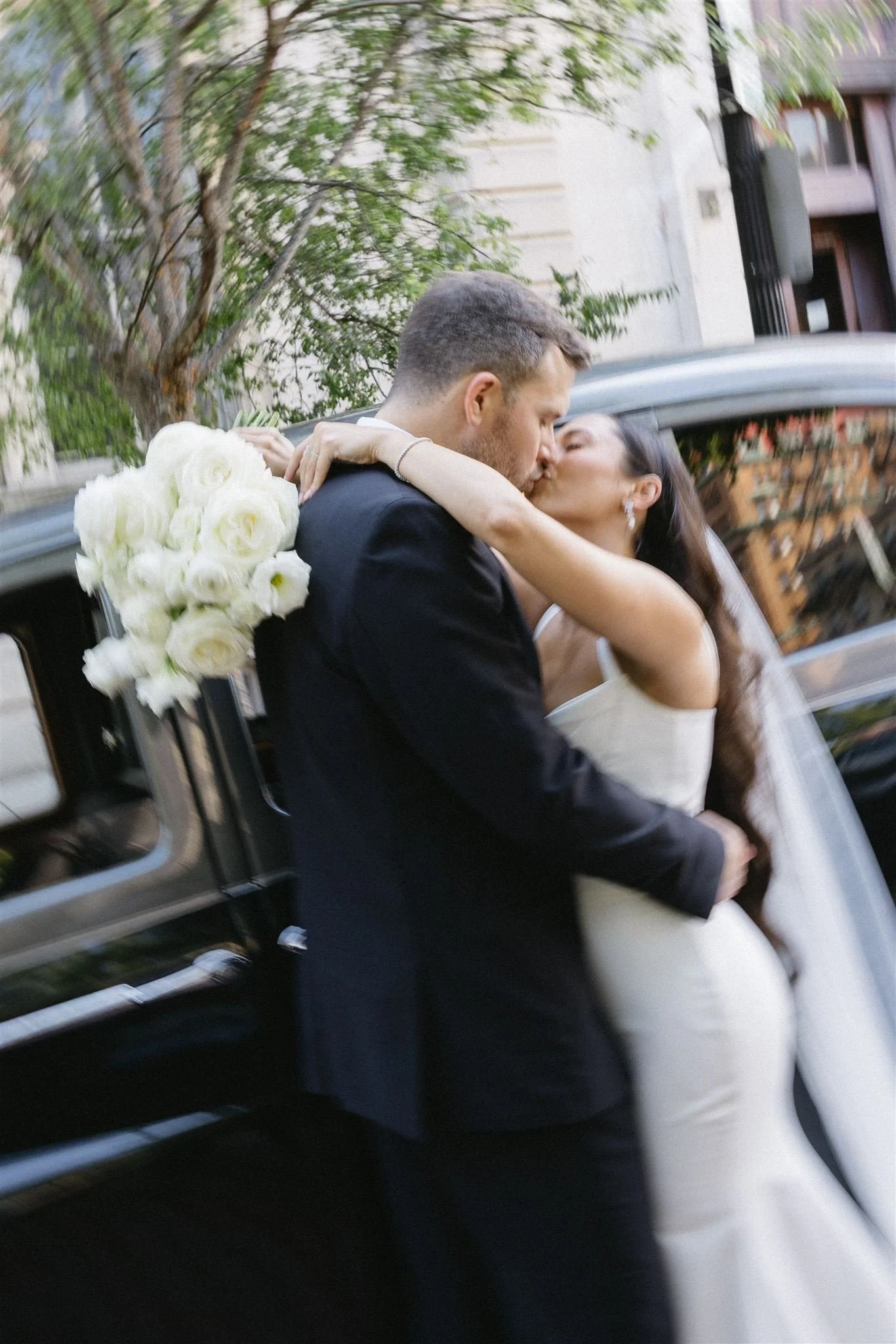 A bride and groom sharing a kiss outside a black car, the bride holding a bouquet of white roses, with trees and buildings in the background.