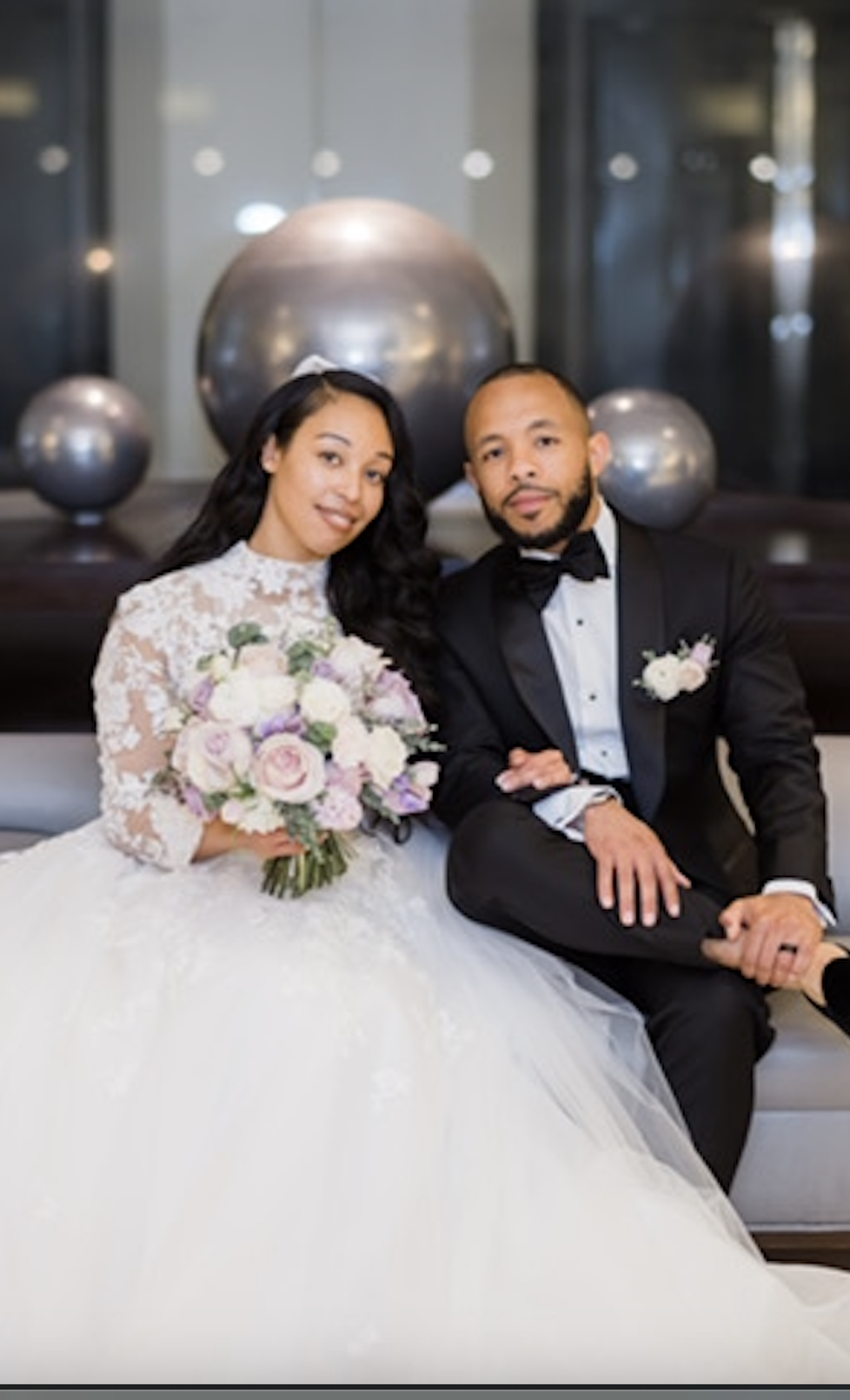 A bride in a white lace wedding dress holding a bouquet of roses and a groom in a black tuxedo sitting on a beige couch with metallic spherical decorations in the background.