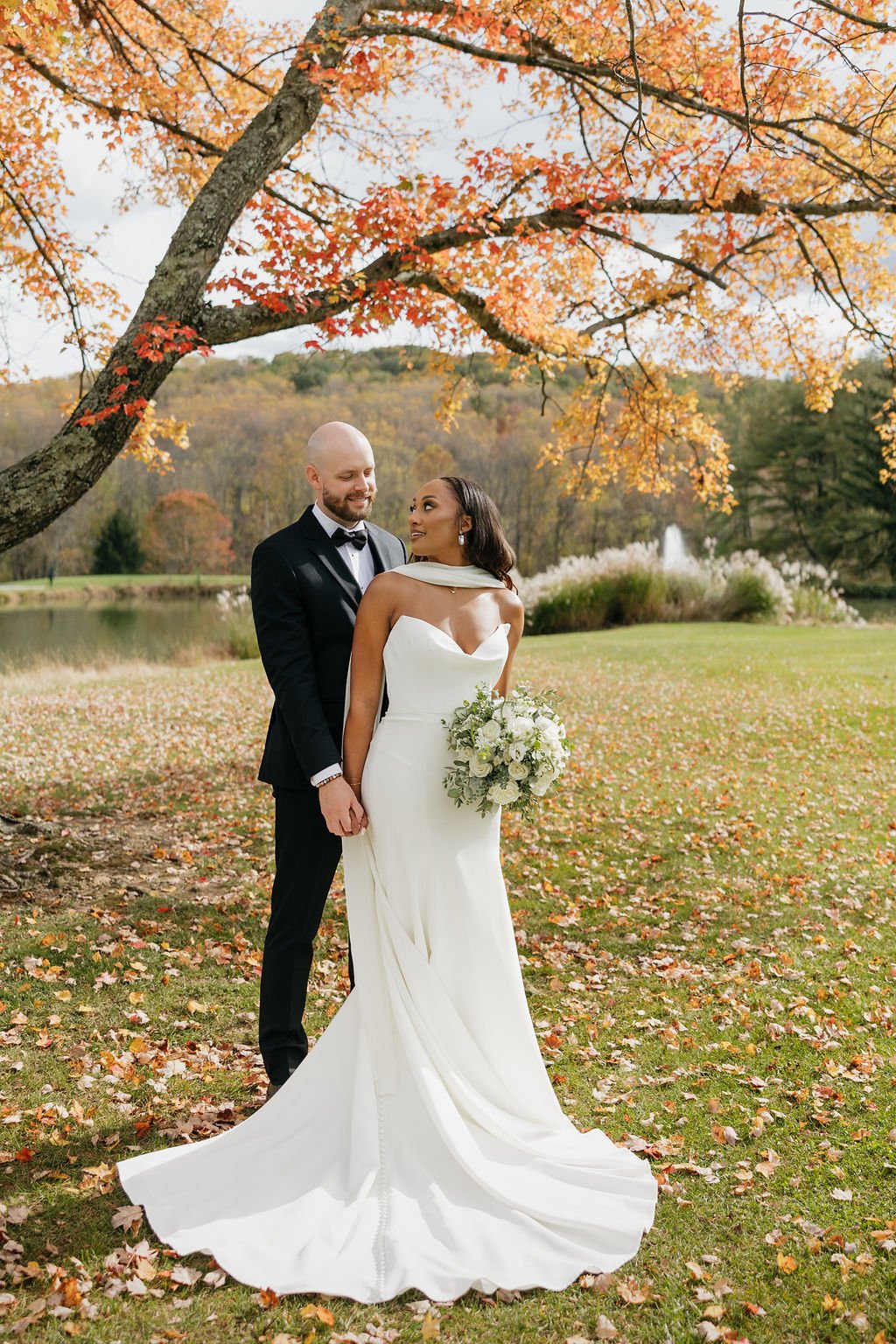 A newlywed couple standing outdoors under an autumn tree, holding hands and gazing at each other. The bride is in a white wedding gown holding a bouquet, and the groom is in a black tuxedo. The scene is set by a lake with a fountain, surrounded by fall foliage.
