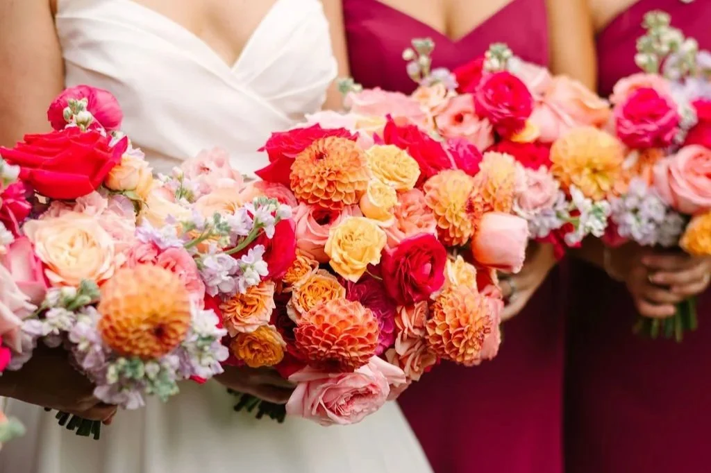 Bridesmaids holding bouquets of colorful flowers, including roses, dahlias, and snapdragons, at a wedding.