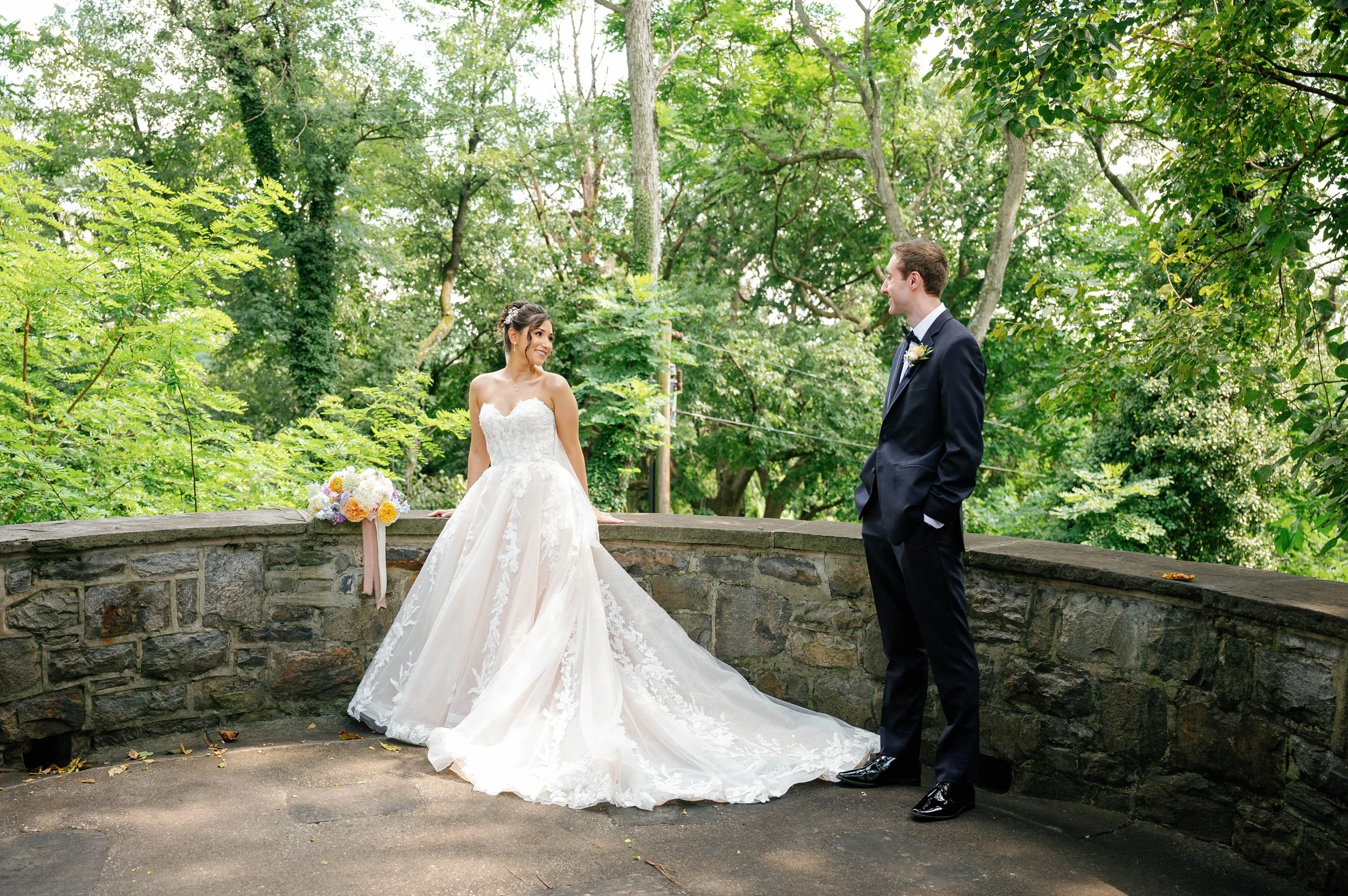 A bride and groom stand outdoors on a stone balcony, surrounded by lush green trees. The bride wears a white lace wedding gown and the groom a black tuxedo. The bride looks at the groom with a smile, and a bouquet of flowers rests on the stone wall beside her.