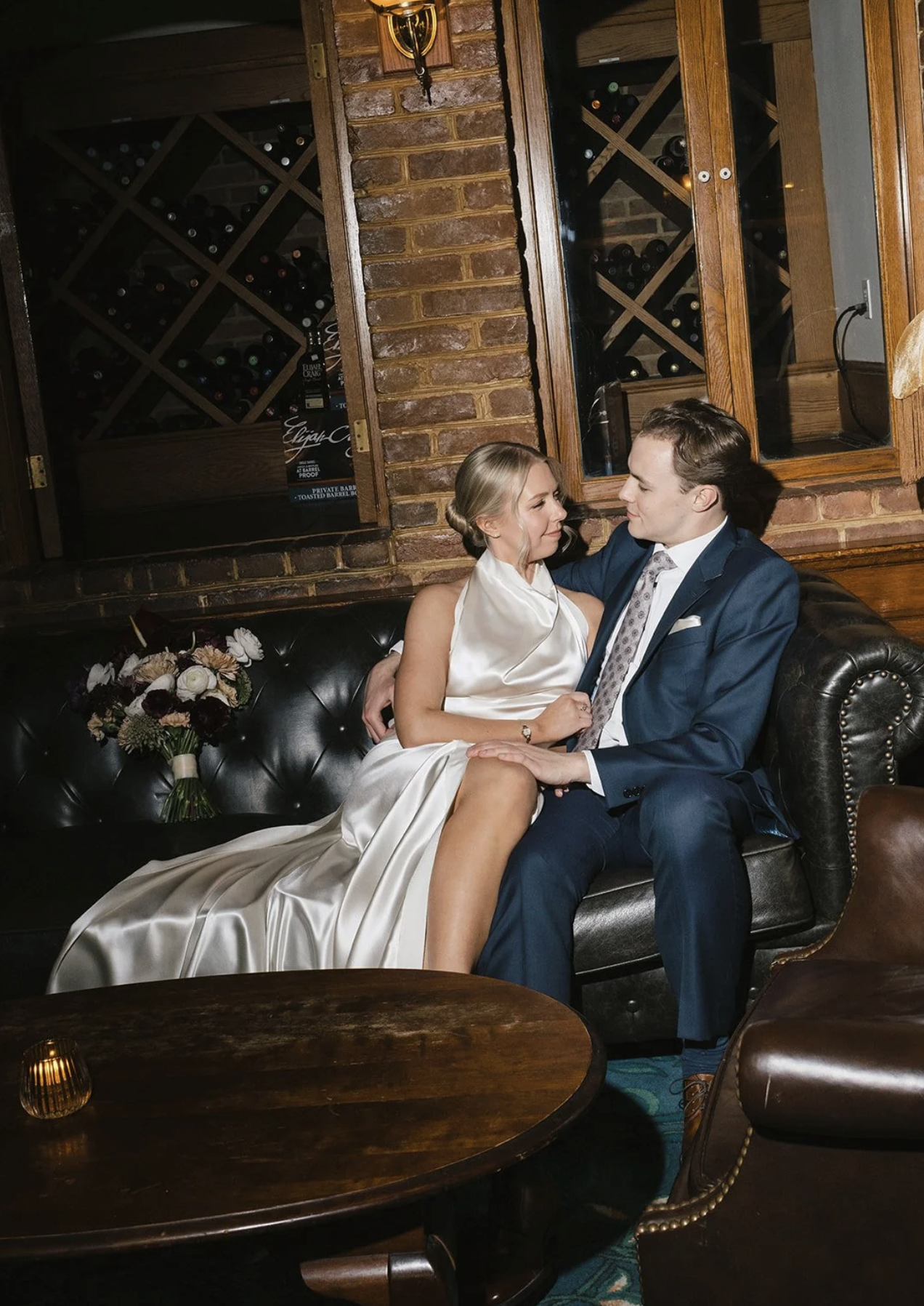 A bride and groom sitting on a black leather sofa in a cozy, rustic room with brick walls and wooden furniture. The bride is in a white satin wedding gown, and the groom is in a navy blue suit. They are gazing at each other affectionately, and a bouquet of flowers rests beside them.