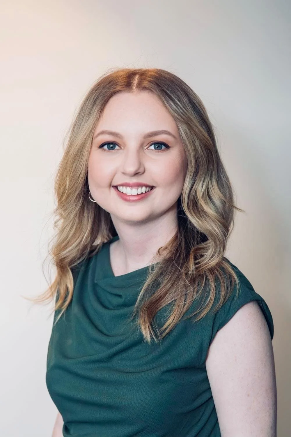 A woman with long wavy blonde hair, blue eyes, and fair skin, smiling and wearing a dark green sleeveless top, standing against a light neutral background.