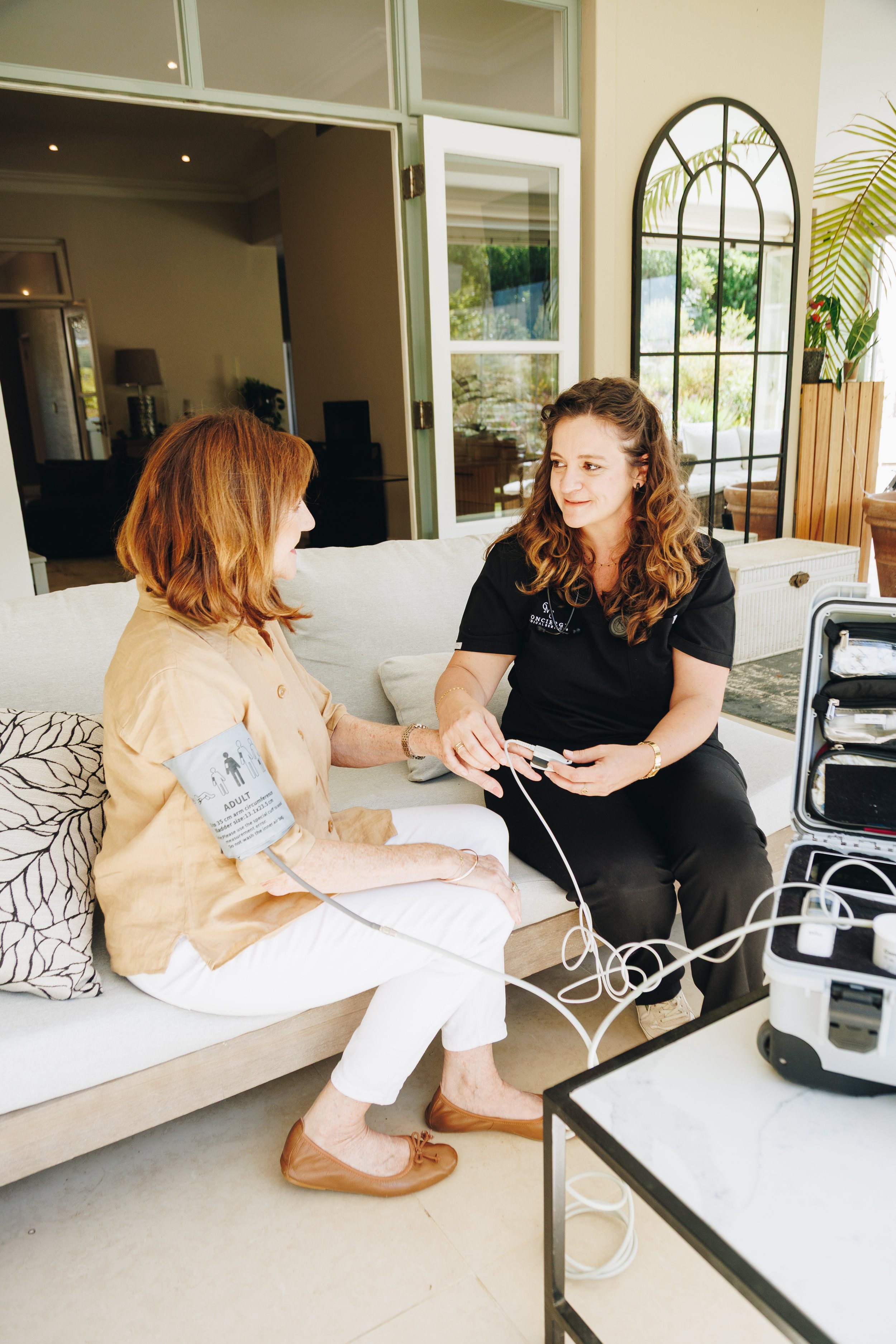 A woman with red hair receiving a blood pressure check from a concierge medical doctor worker with brown curly hair, sitting on a sofa in a bright, modern room with large windows and decorative mirror.