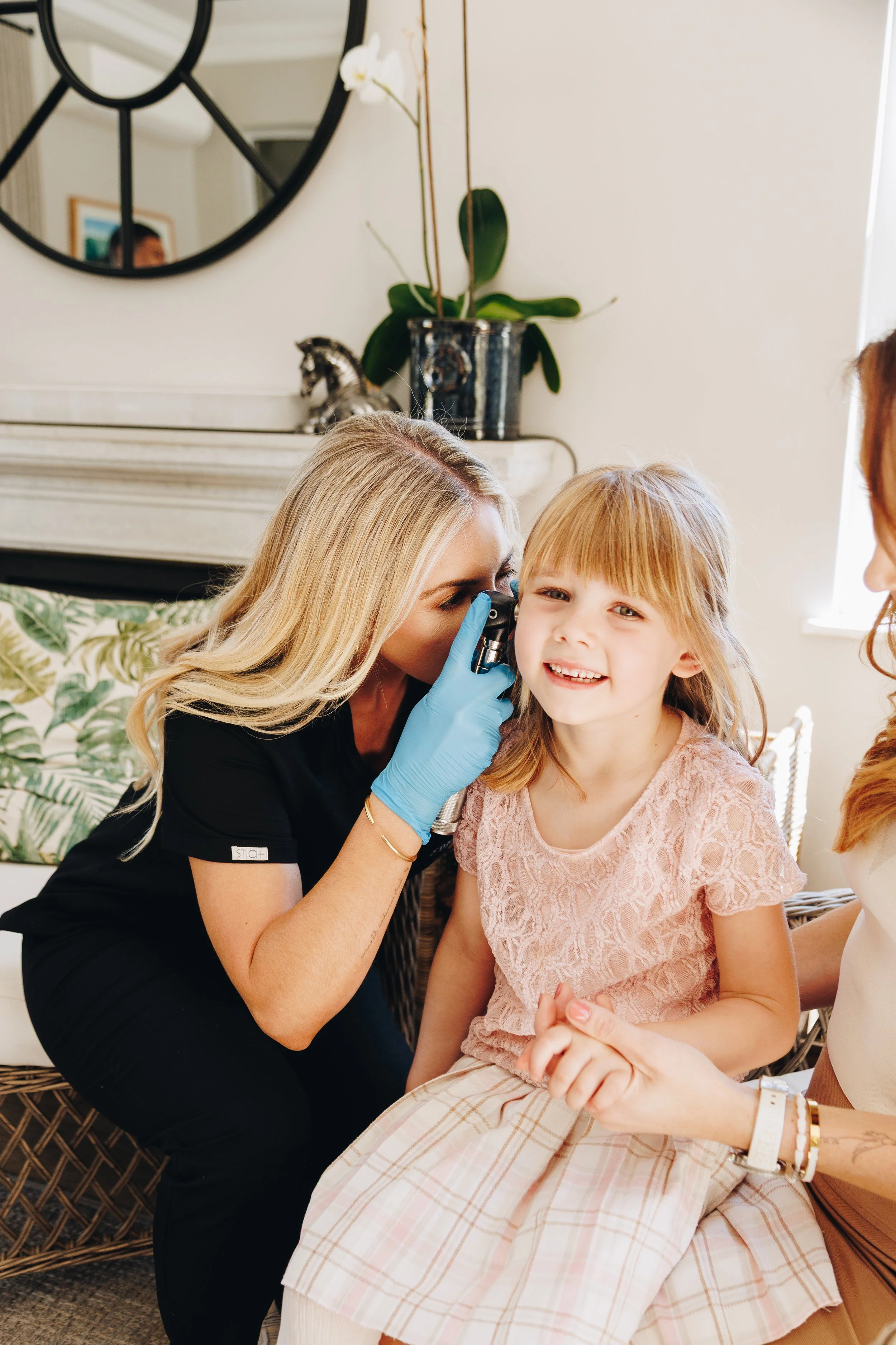 A woman doctor in black scrubs and blue gloves looks into a child's ear with an otoscope, while a young girl with blonde hair and a pink lace top sits on her lap, smiling. Another woman with red hair is partially visible holding the girl.