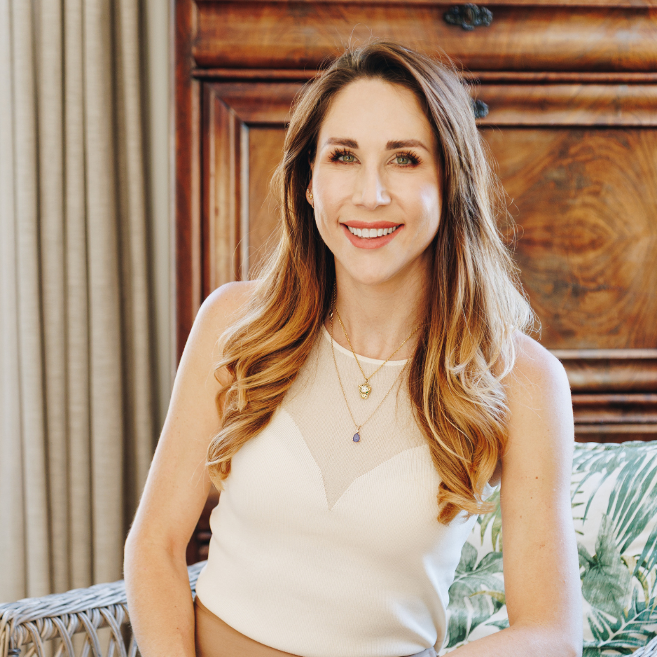A smiling woman with long wavy hair in a sleeveless cream top with a mesh neckline, sitting in a room with wooden furniture and curtains.