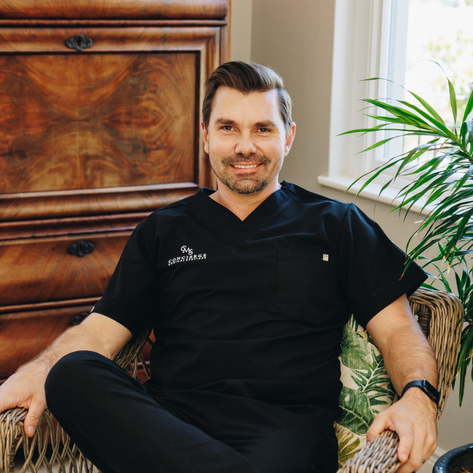 A smiling male medical doctor with dark hair and a beard, dressed in a black medical uniform, sitting in a wicker chair indoors near a window with green plants.