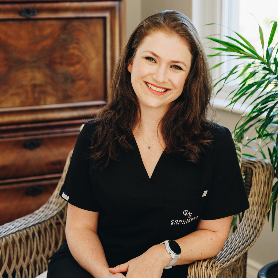 A smiling woman medical doctor with long brown hair wearing a black medical uniform and sitting in a wicker chair in a room with a wooden dresser and a green potted plant.