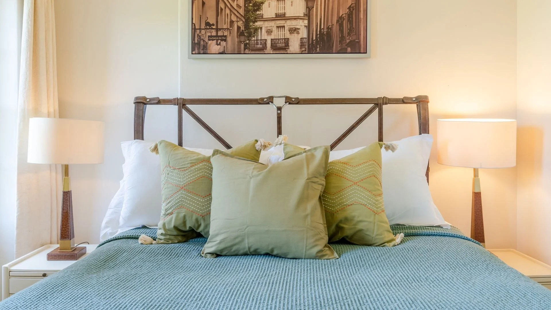 Cozy bed with green pillows, blue textured blanket, and two bedside lamps in a softly lit room.