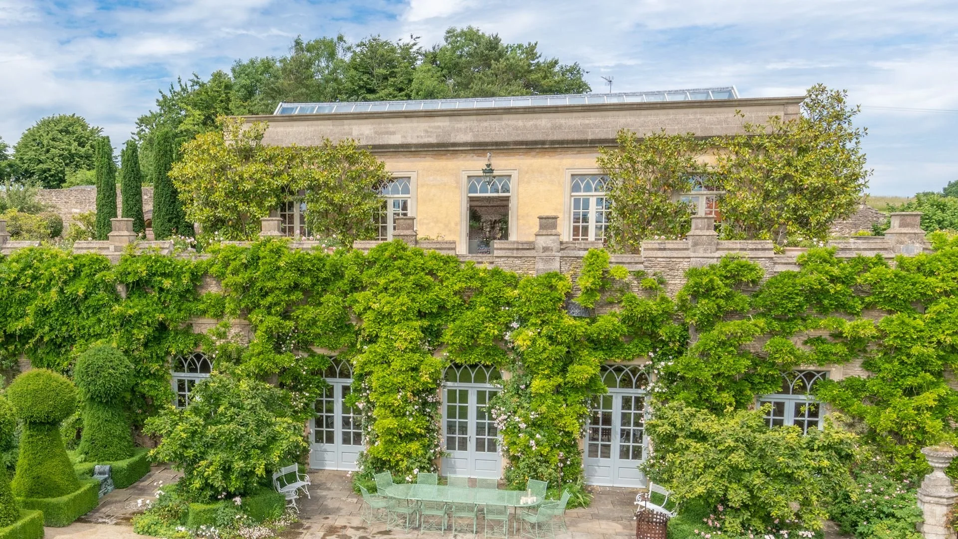 Large stone estate with ivy-covered walls and arched windows, surrounded by lush gardens and topiary. Outdoor seating includes a green metal table and chairs on a stone patio.