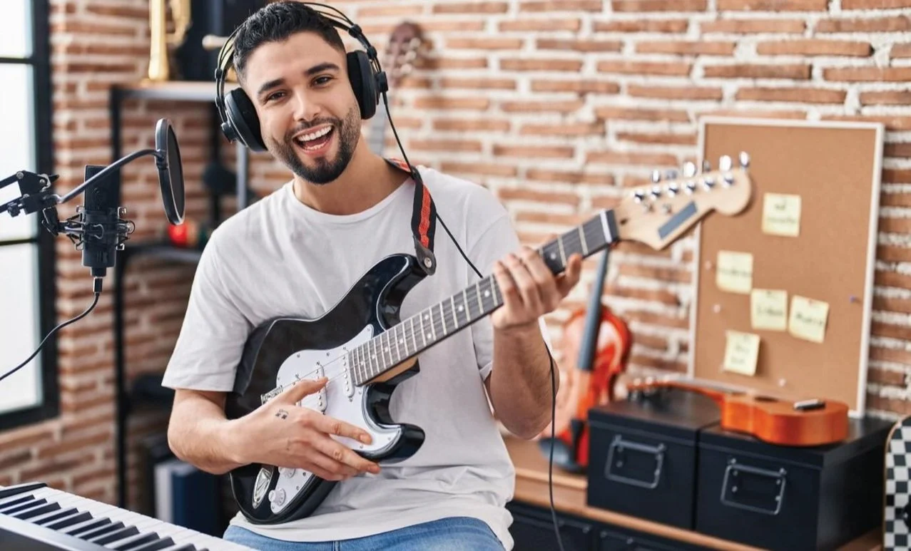 Man with headphones playing electric guitar next to a microphone in a music studio.