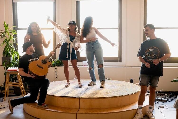 A group of five people enjoying a casual performance; two women sing and dance on a round wooden platform, one man plays an acoustic guitar, and two others appear to be enjoying the music.