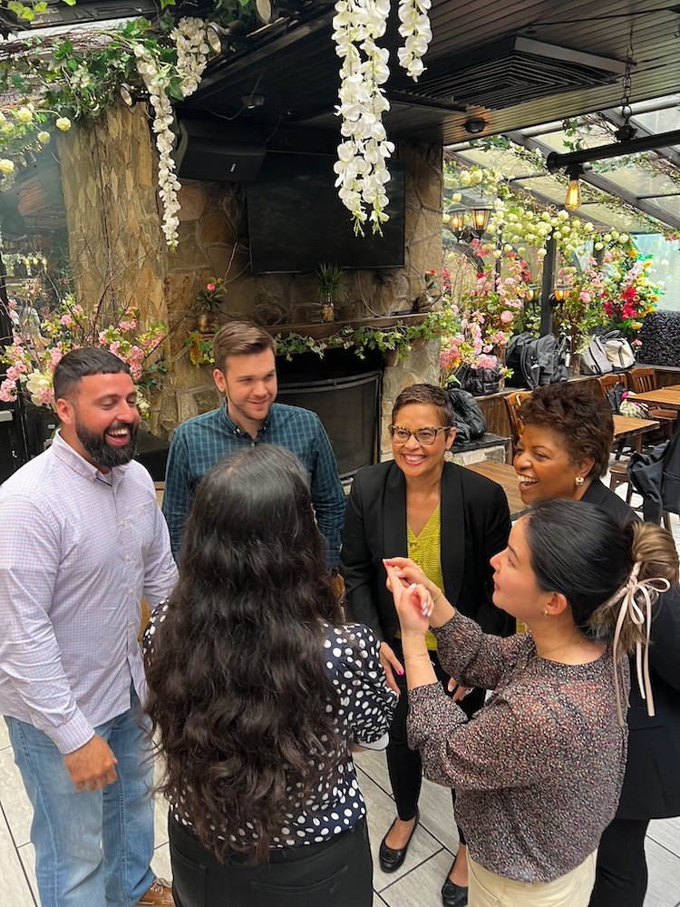 A group of six people, four women and two men, standing in a circle and conversing in a bright, floral-decorated setting. The atmosphere is friendly and lively, with hanging white flowers and colorful plants in the background, creating a garden-like