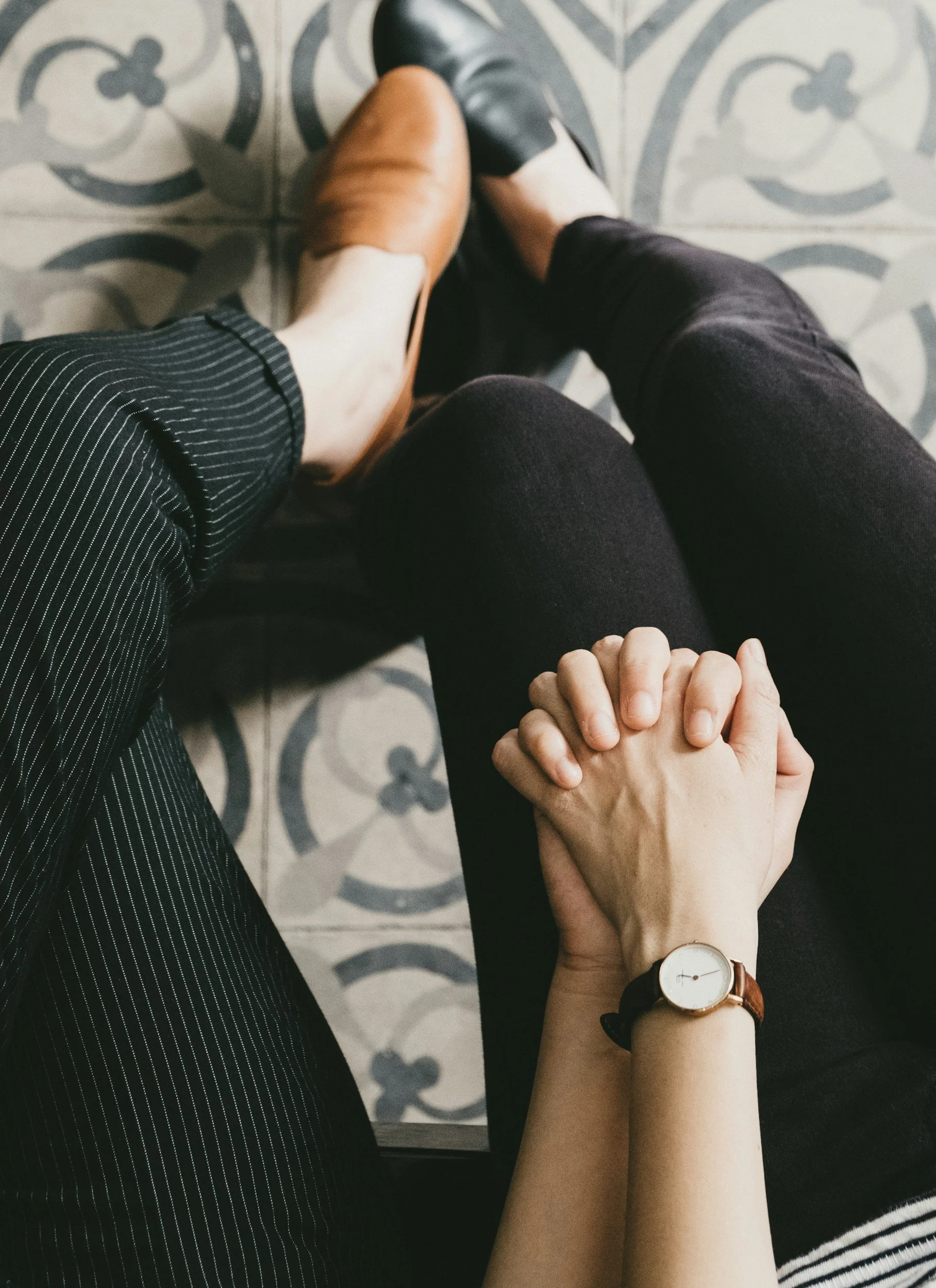 Couple sitting on patterned floor, holding hands, one wearing a watch, one barefoot.
