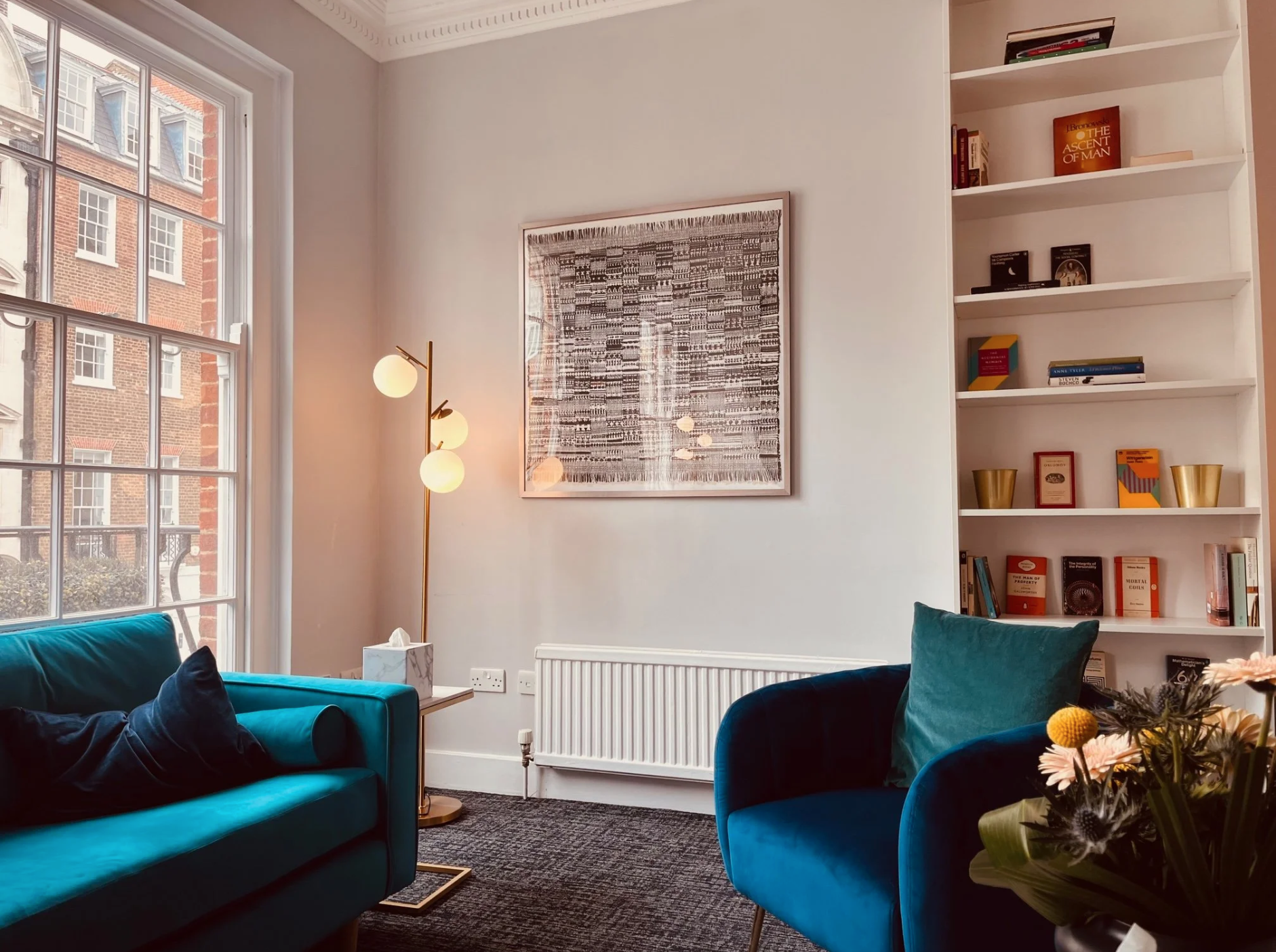 A cozy living room with a large window, teal velvet sofa with a pillow, a black armchair with a teal pillow, a black rug, a tall gold floor lamp with round white bulbs, a white bookshelf with books and decorative items, a framed abstract art piece on the white wall, a white radiator, a small side table with tissues, and a bouquet of flowers in the foreground.