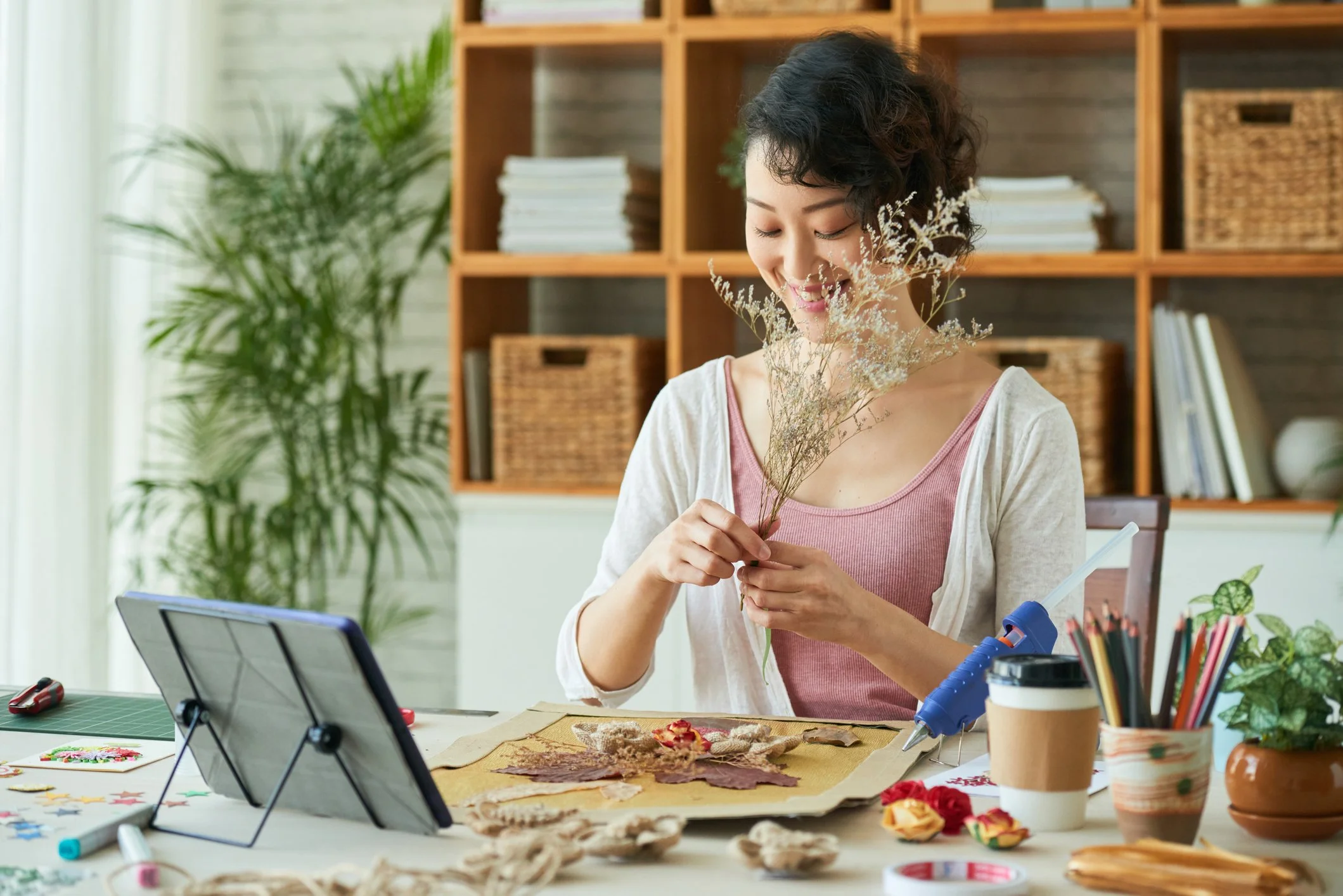 A woman with short curly black hair smiling while arranging dried flowers on a craft project, surrounded by art supplies and decorative items on a table in a cozy room.