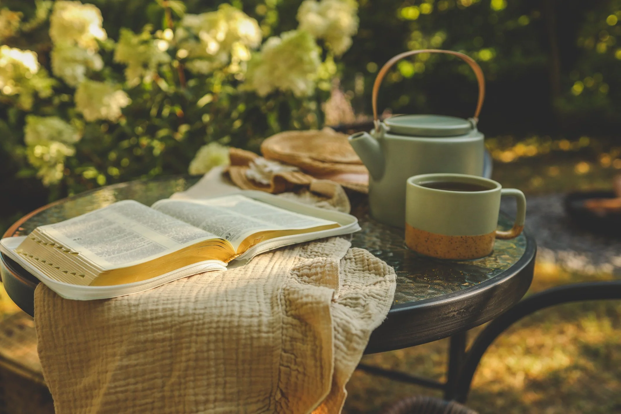 A table with an open Bible, a yellow notepad, a cream-colored napkin, a light green teapot, a mug of coffee, and a small plate, set outdoors with green foliage in the background.