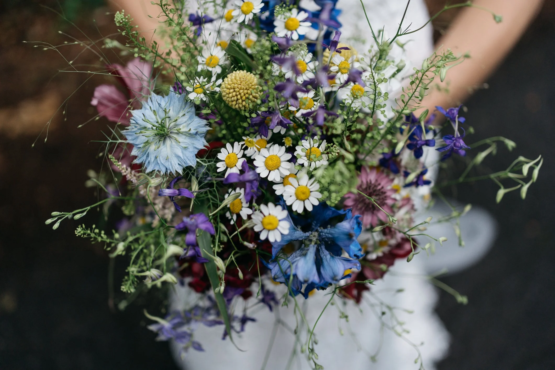 A close-up of a vibrant mixed flower bouquet including daisies, purple bellflowers, blue delphiniums, and yellow billy balls.