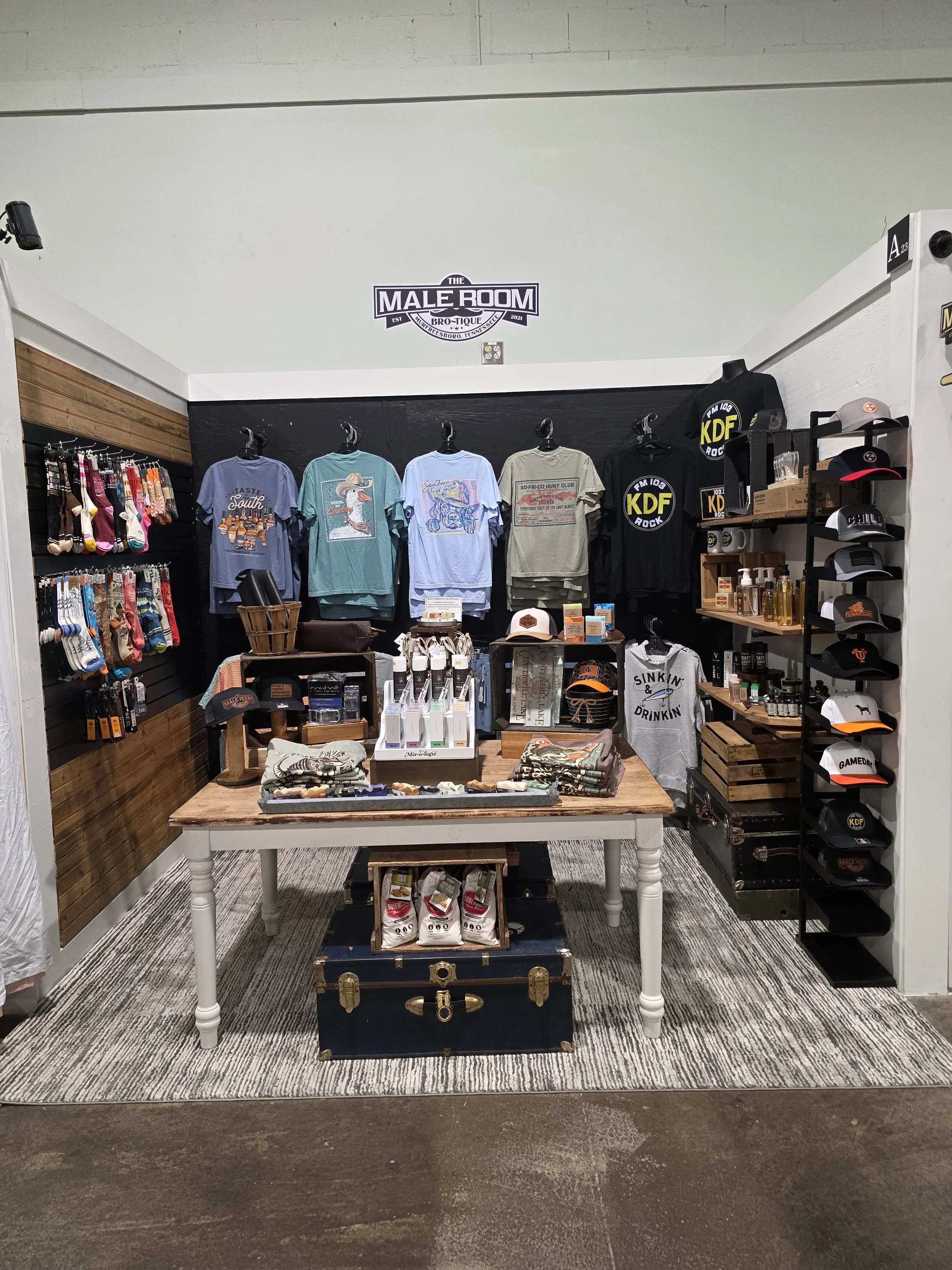 The Male Room Brotique Booth A-23 display inside The Collective Boutiques in Murfreesboro, Tennessee, featuring graphic tees, snapback trucker hats, novelty socks, and men's grooming products.